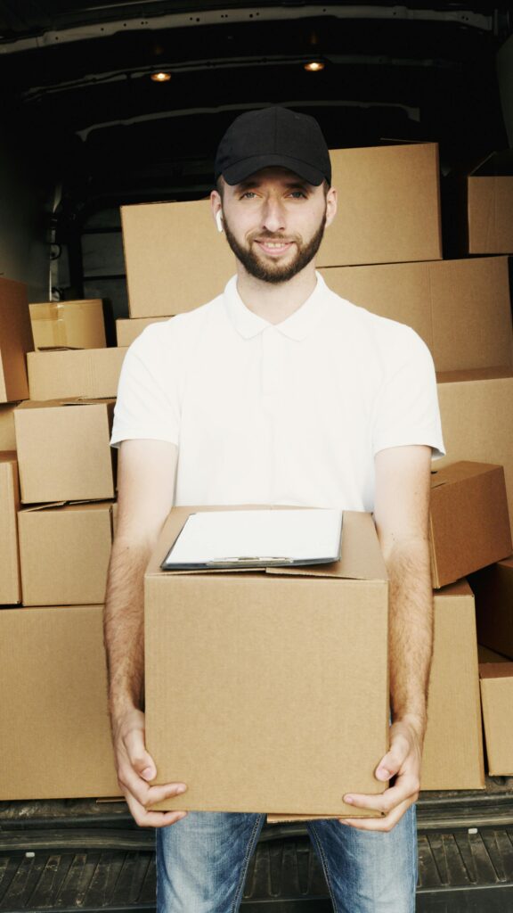 pexels photo 5025483 5025483 A deliveryman in a cap holding a box standing in front of a truck loaded with cartons.