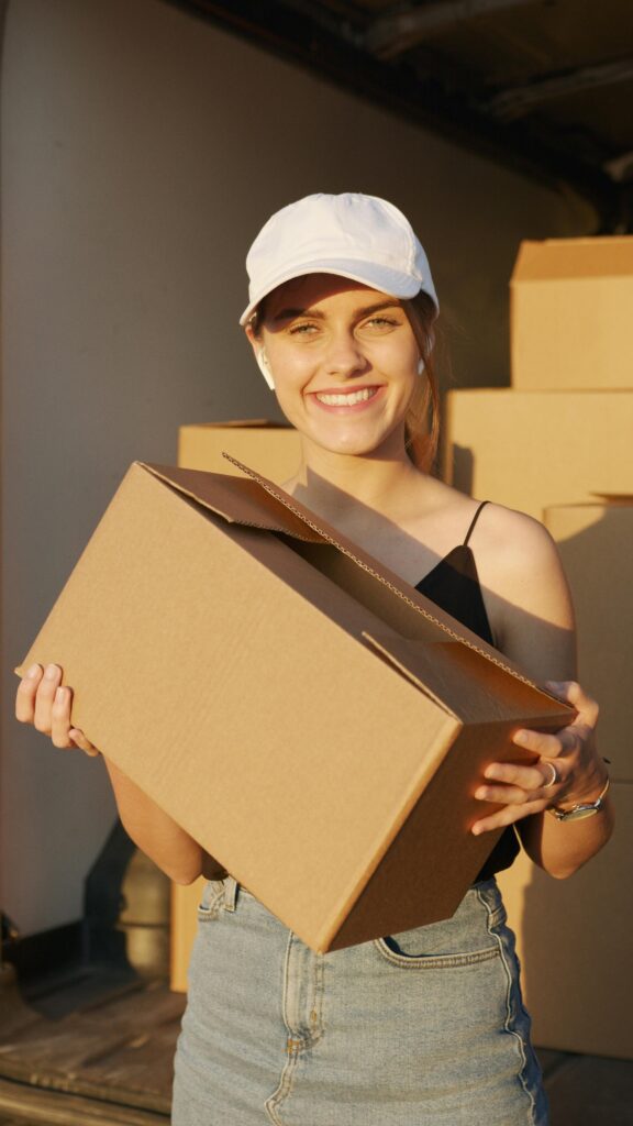 pexels photo 5025510 5025510 A cheerful woman wearing a cap holds a cardboard box with sunlight illuminating her face.