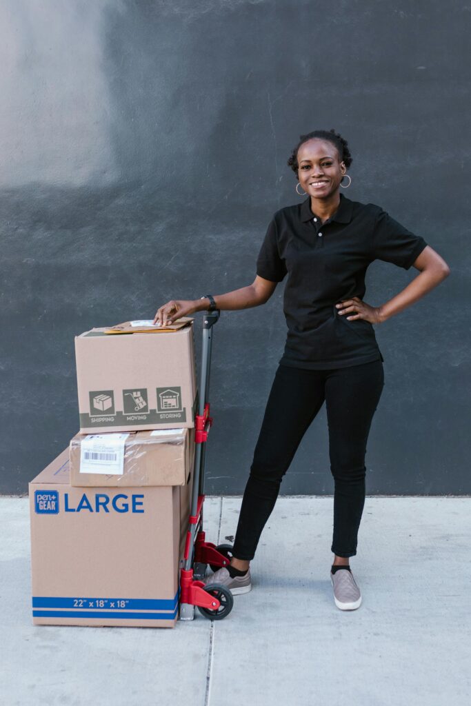 pexels photo 7362931 7362931 Smiling woman with boxes on a trolley cart, ideal for delivery service themes.